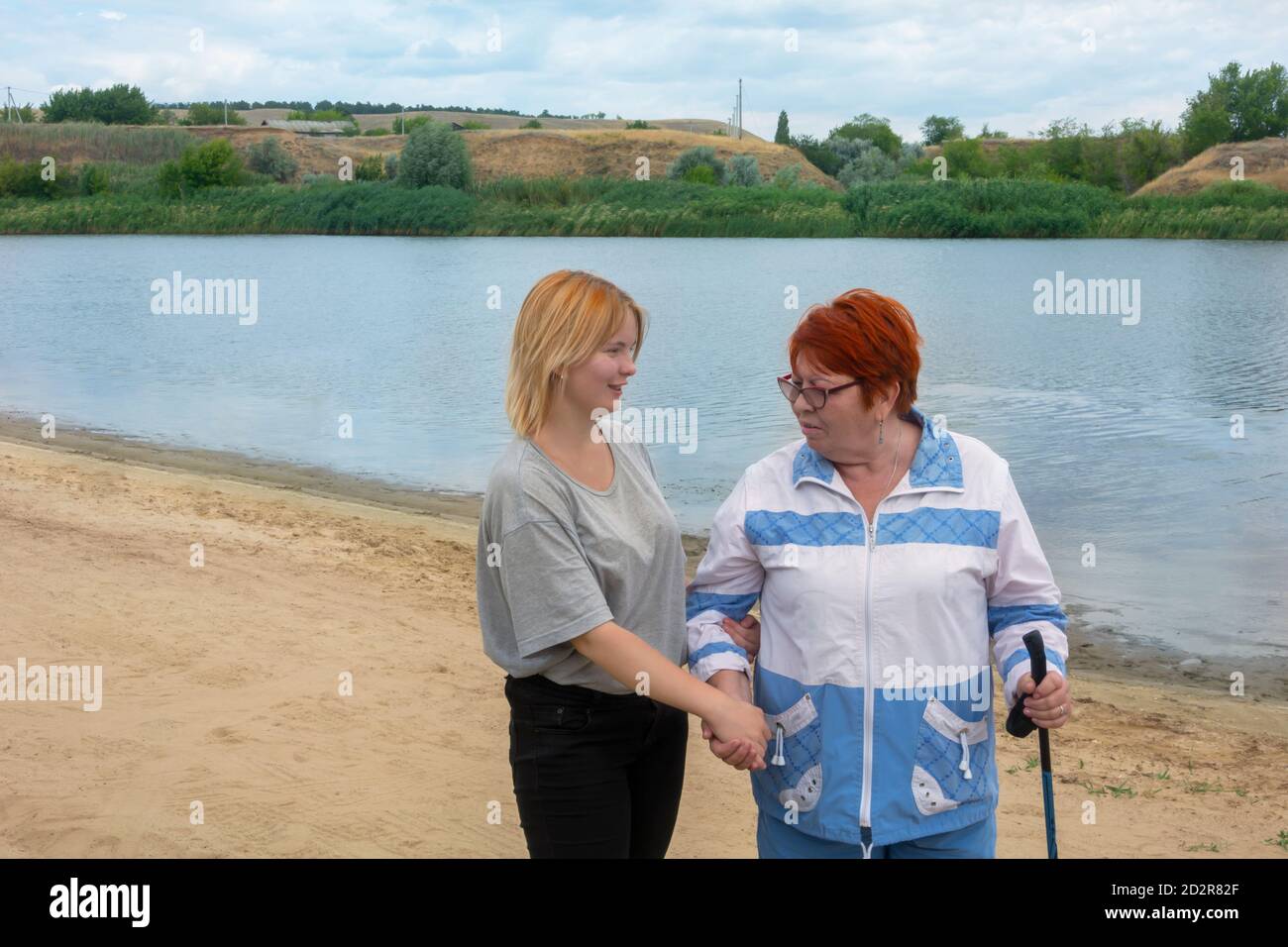 La nipote aiuta la nonna a camminare lungo il fiume, la sostiene a mano. Il concetto di comunicazione tra generazioni, il rispetto per gli anziani. Foto Stock