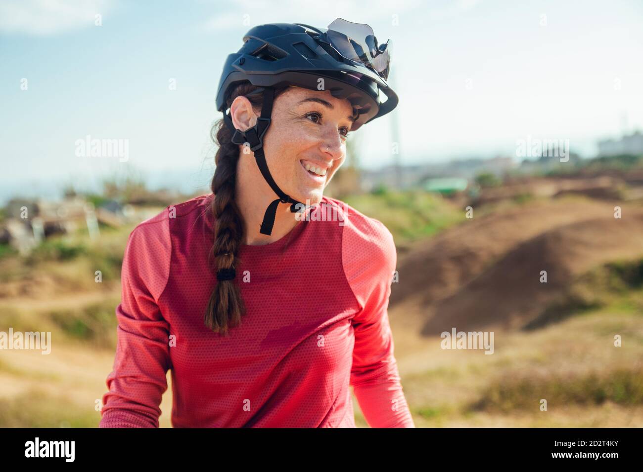 Sportswoman con casco nero e abbigliamento sportivo rosso con occhiali che riposano vicino alla pista da allenamento guardando lontano Foto Stock