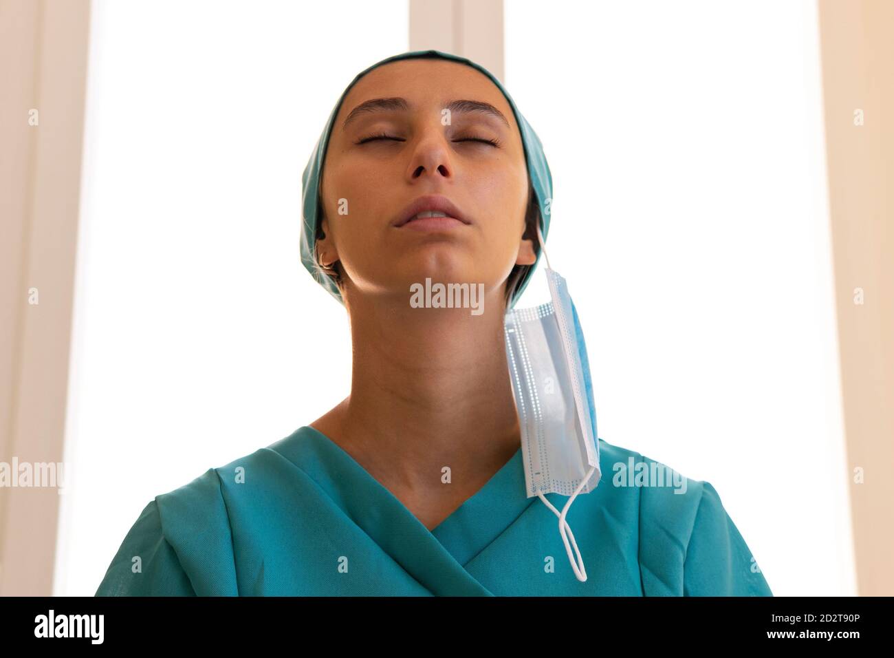 Basso angolo di tranquilla medica femminile in uniforme in piedi dentro clinica senza maschera e respirazione in aria fresca con chiuso occhi dopo l'epidemia di coronavirus Foto Stock