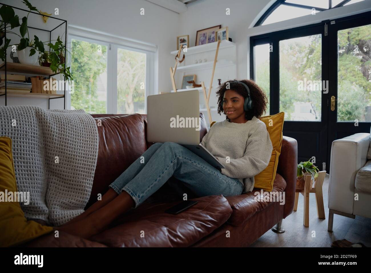 Donna seduta sul divano utilizzando un computer portatile per guardare video con le cuffie a casa Foto Stock