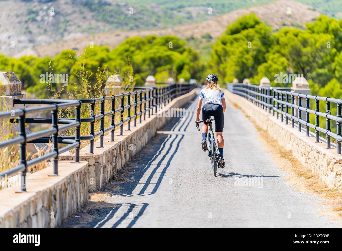 Vista posteriore completa del ciclista FIT femmina in abbigliamento sportivo e casco in bicicletta su strada curva stretta che conduce attraverso verde foresta in estate giorno Foto Stock