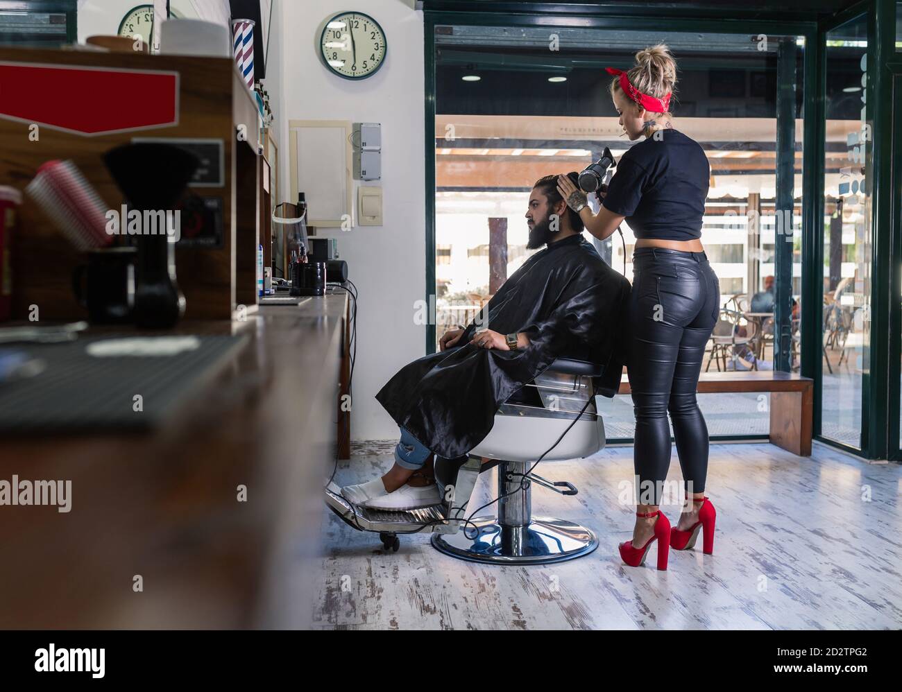 Vista laterale dell'elegante master femminile in pantaloni in pelle nera e tacchi alti rossi che asciugano i capelli del cliente maschio bearded mentre si lavora in barbiere Foto Stock