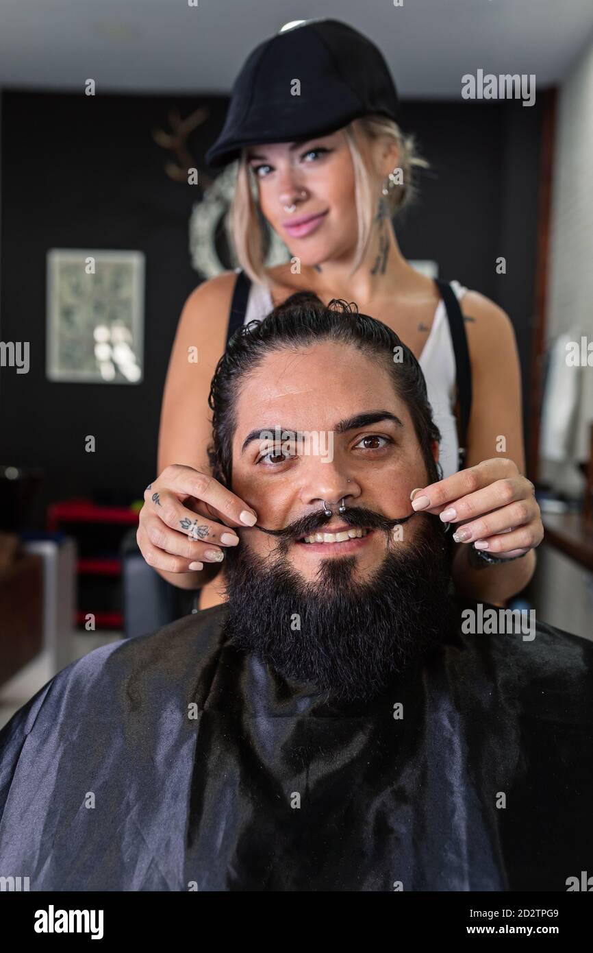 Soddisfatto cliente di sesso maschile sorridente con barba e baffi seduti in sedia con il barbiere femminile in piedi dietro e guardando fotocamera Foto Stock