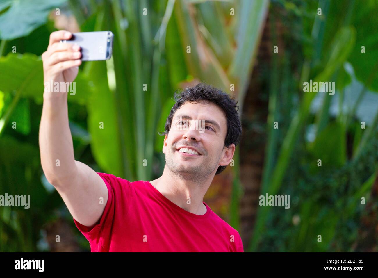 Giovane uomo con smartphone che fa un selfie, vestito con t-shirt rossa su sfondo verde naturale alla stazione ferroviaria Foto Stock