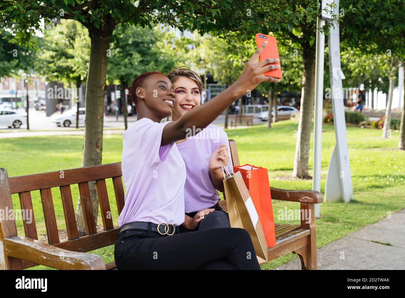 Felice giovane studentesse multirazziali di stile in abiti simili seduti in panchina con borse per la spesa e presa di selfie sullo smartphone mentre si rilassa insieme nel parco dopo il tempo di shopping Foto Stock