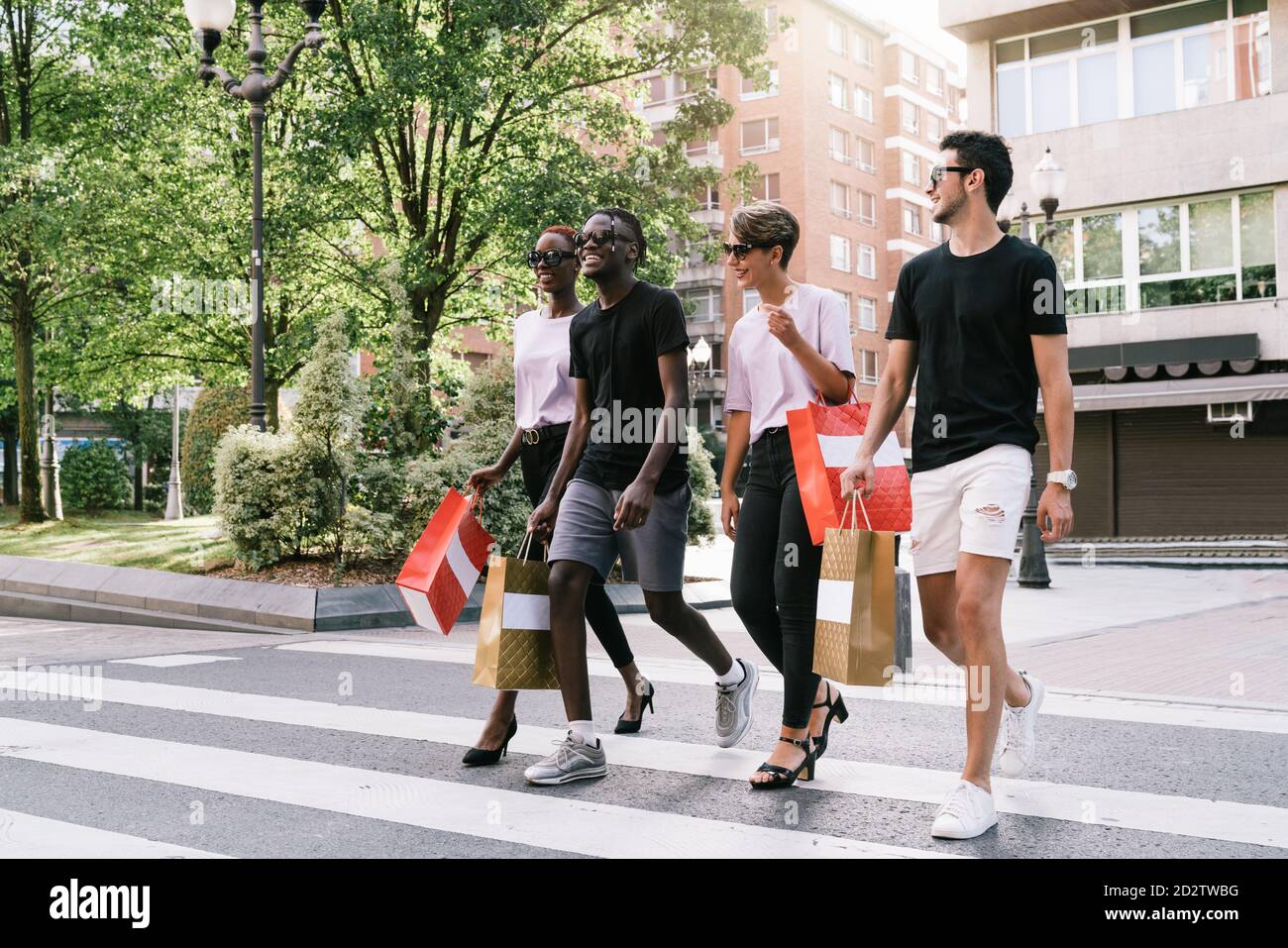 Una lunga serie di giovani studenti allegri e diversi in abiti alla moda e occhiali da sole che trasportano gli acquisti nelle borse di shopping e chiacchierando felicemente mentre si attraversa la strada sulla strada della città Foto Stock