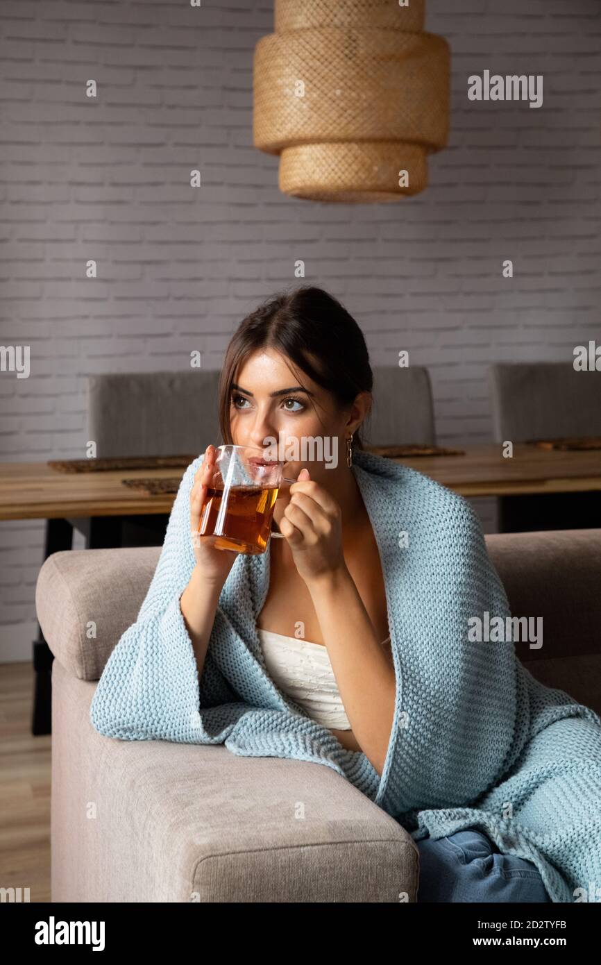 Giovane donna pusiva avvolta in coperta calda seduta sul divano e gustando il tè caldo mentre si rilassa nell'accogliente soggiorno a casa Foto Stock