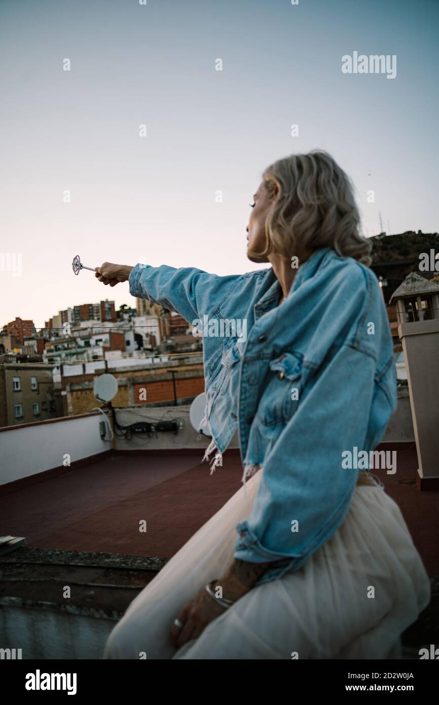Vista laterale di una donna sognante seduta sul tetto dell'edificio e lanciare incantesimo con bacchetta magica in mano estesa mentre guardando via Foto Stock