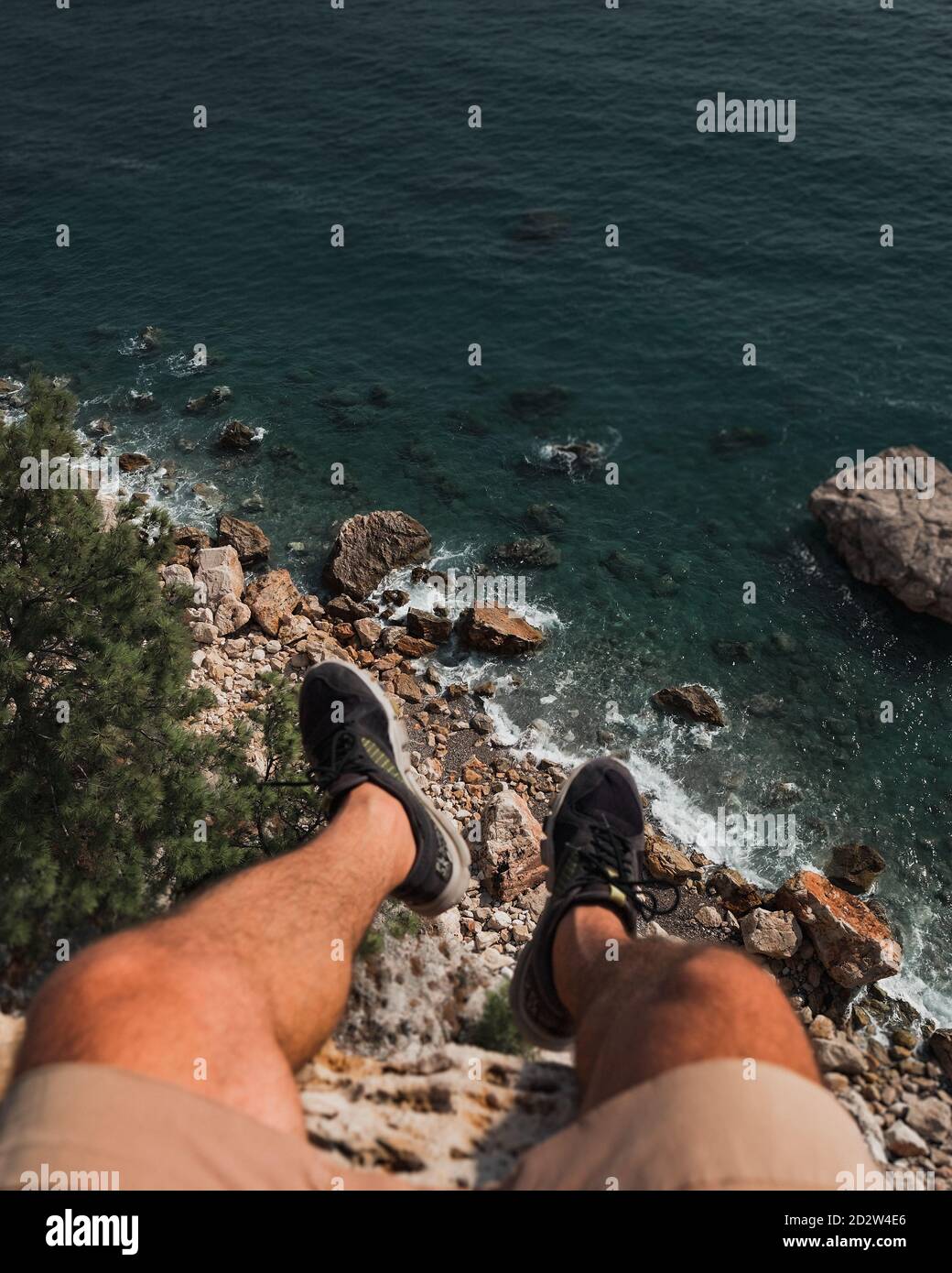 Le gambe dell'uomo sono appese dal bordo della scogliera sopra il mare e le rocce. Foto Stock