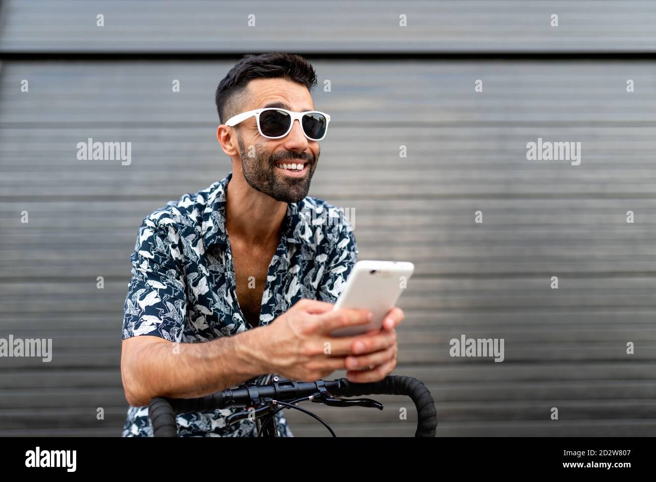 Un uomo che si è deliziato con un abito elegante appoggiato sul manubrio della bicicletta e leggere messaggi sui social media tramite telefono cellulare in città Foto Stock
