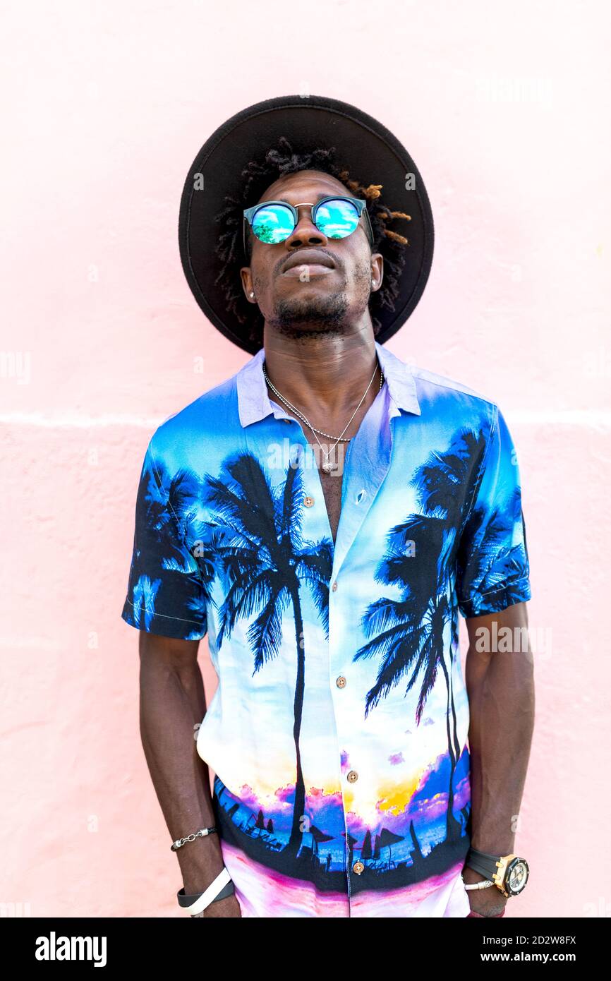 Uomo afro-americano sicuro che indossa una camicia trendy con palme ed eleganti occhiali da sole in piedi sulla strada vicino all'edificio con rosa parete Foto Stock