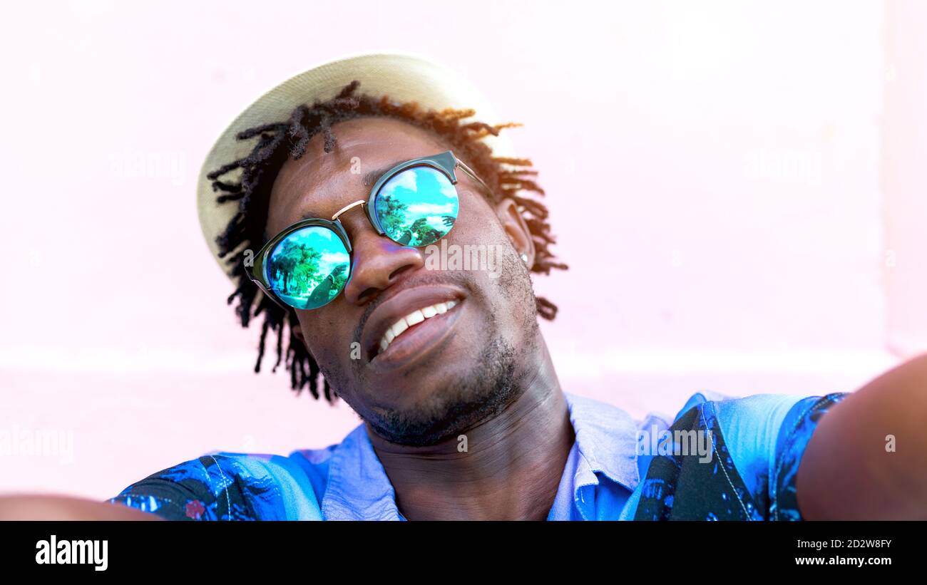 Uomo etnico che indossa una camicia elegante con stampa tropicale in piedi vicino alla costruzione in città e prendendo selfie su smartphone Foto Stock