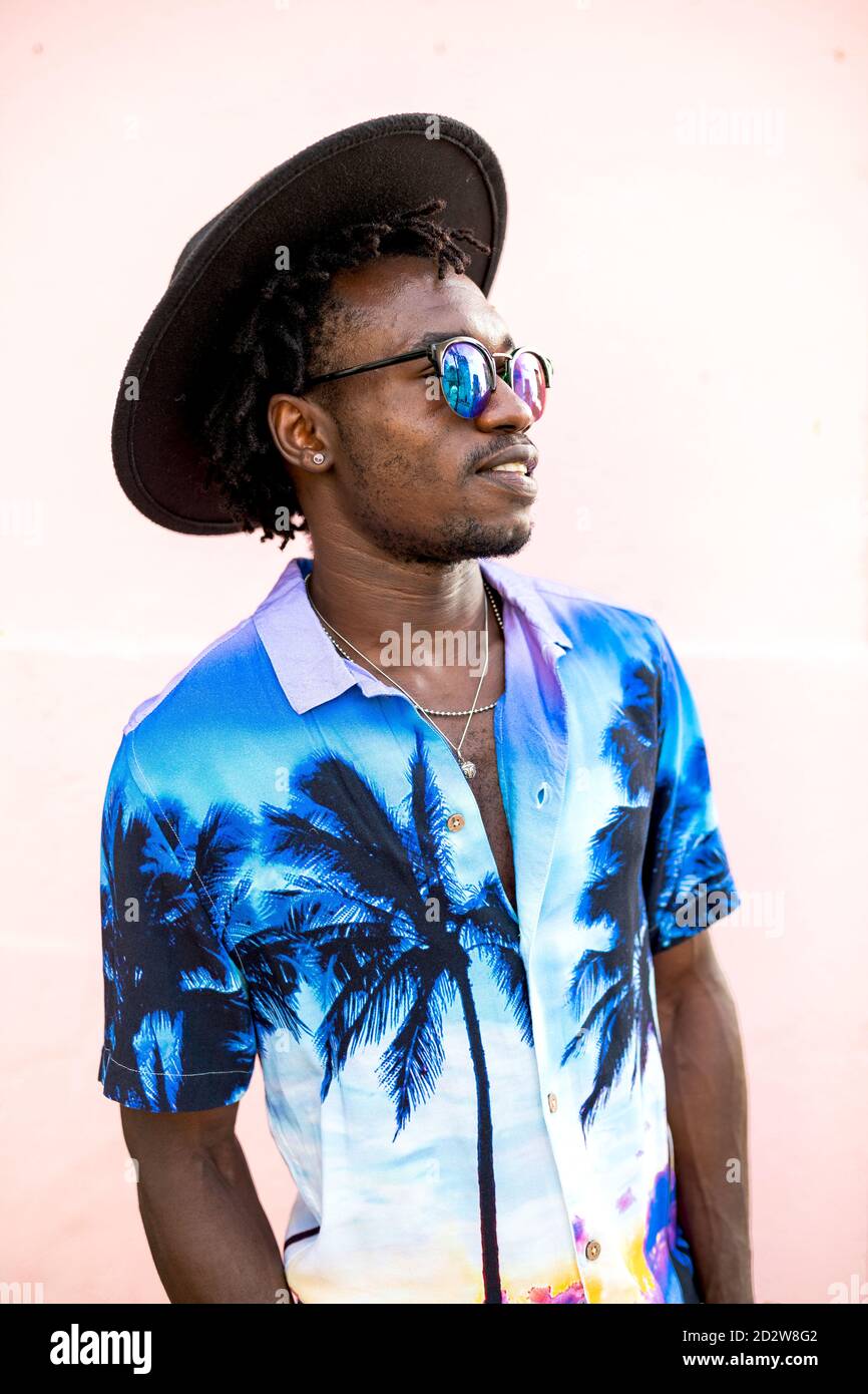 Uomo afro-americano sicuro che indossa una camicia trendy con palme, cappello e occhiali da sole eleganti in piedi sulla strada vicino a un edificio con parete rosa Foto Stock