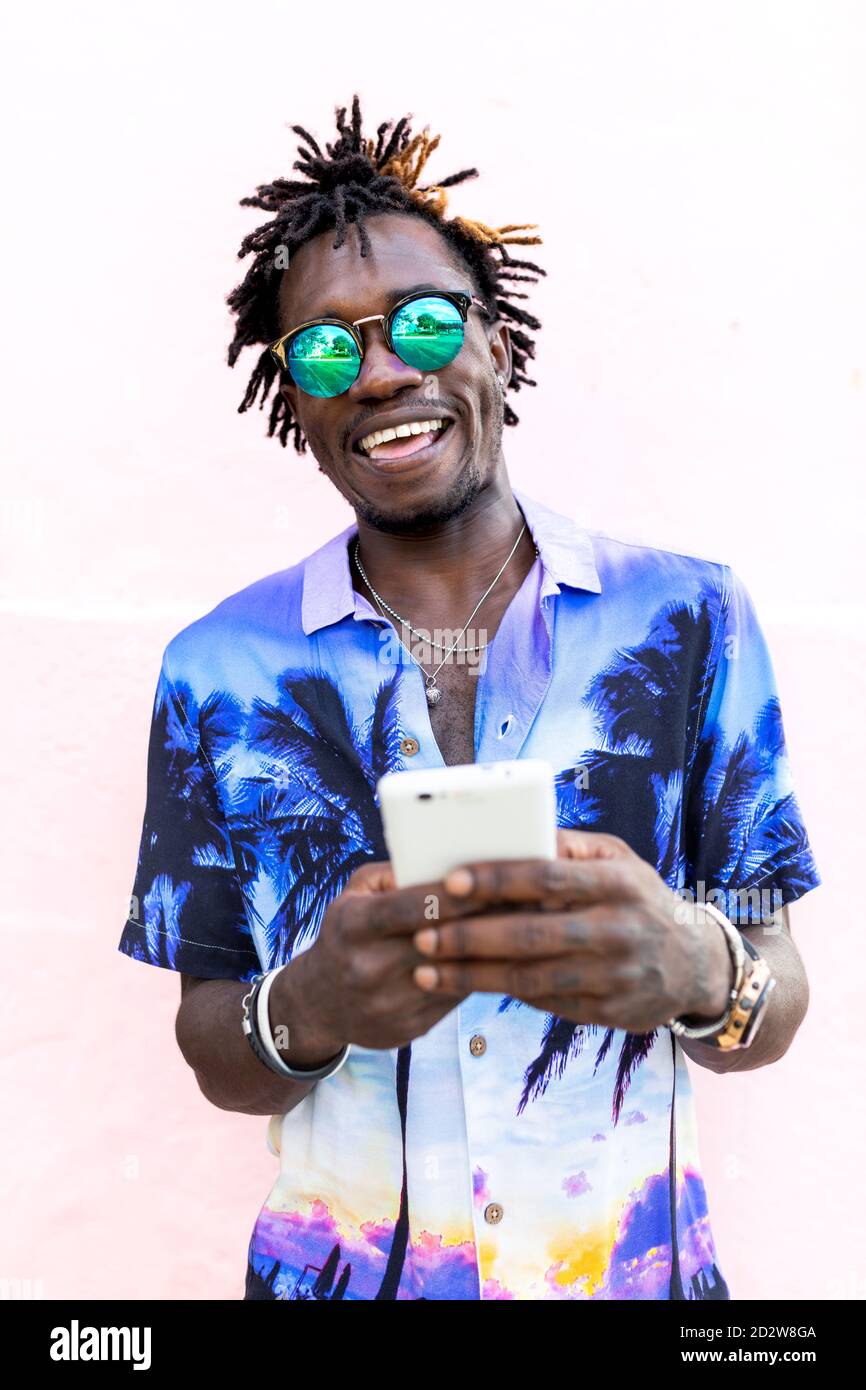 Allegro e trendy uomo afro-americano in camicia con stampa tropicale e con i dreadlock in piedi vicino a un edificio rosa e chiacchierare social media tramite telefono cellulare Foto Stock