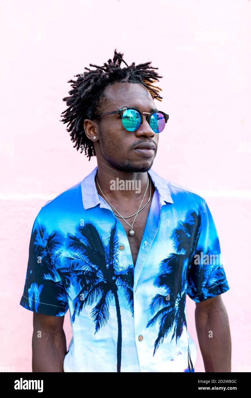 Uomo afro-americano sicuro che indossa una camicia trendy con palme ed eleganti occhiali da sole in piedi sulla strada vicino all'edificio con rosa parete Foto Stock