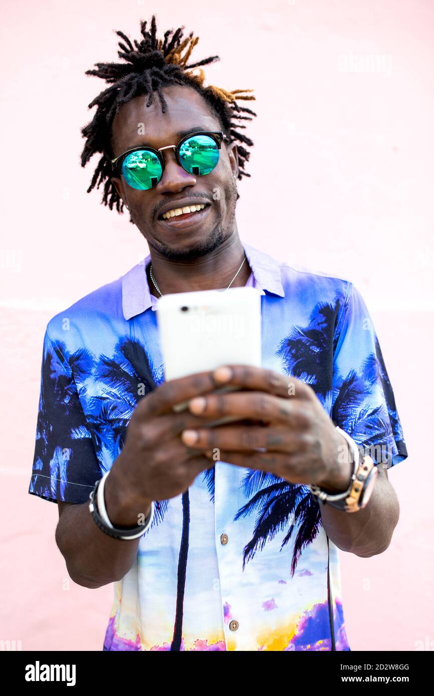 Uomo afro-americano trendy in camicia con stampa tropicale e. con i dreadlock in piedi vicino a un edificio rosa e chiacchierando sui social contenuti multimediali tramite telefono cellulare Foto Stock