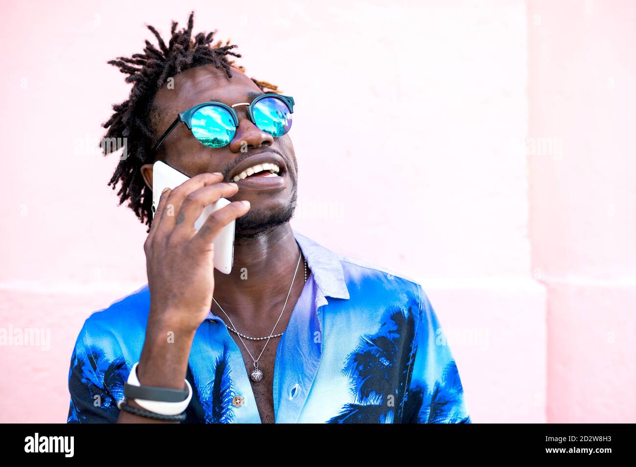 Uomo afro-americano trendy in camicia con stampa tropicale e. con i dreadlock in piedi vicino a un edificio rosa e parlando su un cellulare telefono Foto Stock