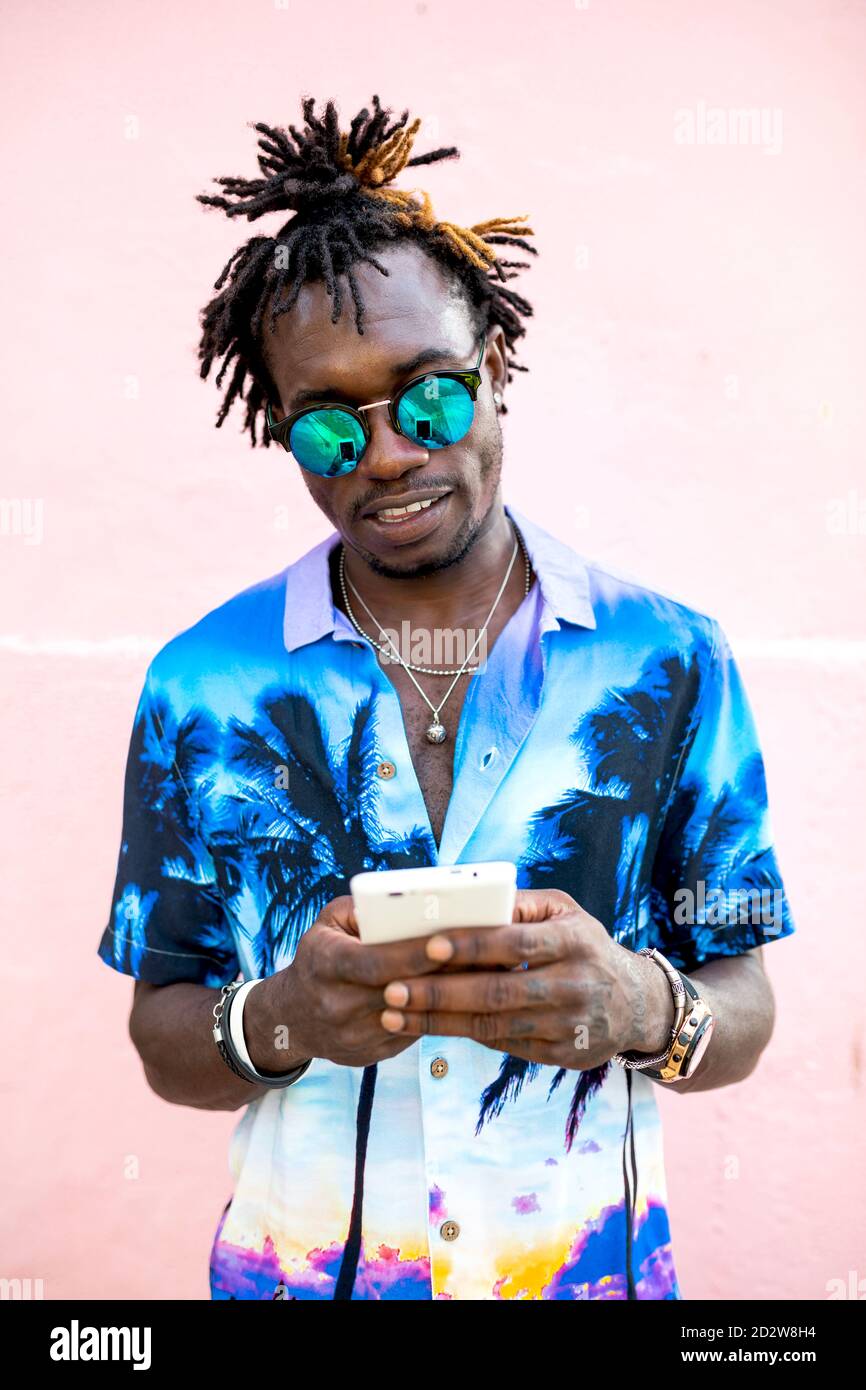 Uomo afro-americano trendy in camicia con stampa tropicale e. con i dreadlock in piedi vicino a un edificio rosa e chiacchierando sui social contenuti multimediali tramite telefono cellulare Foto Stock