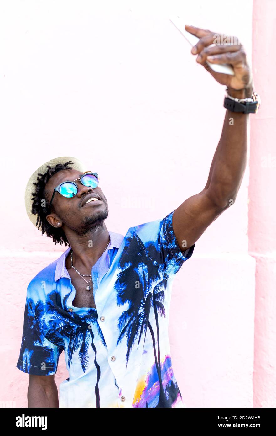 Uomo etnico che indossa una camicia elegante con stampa tropicale in piedi vicino alla costruzione in città e prendendo selfie su smartphone Foto Stock