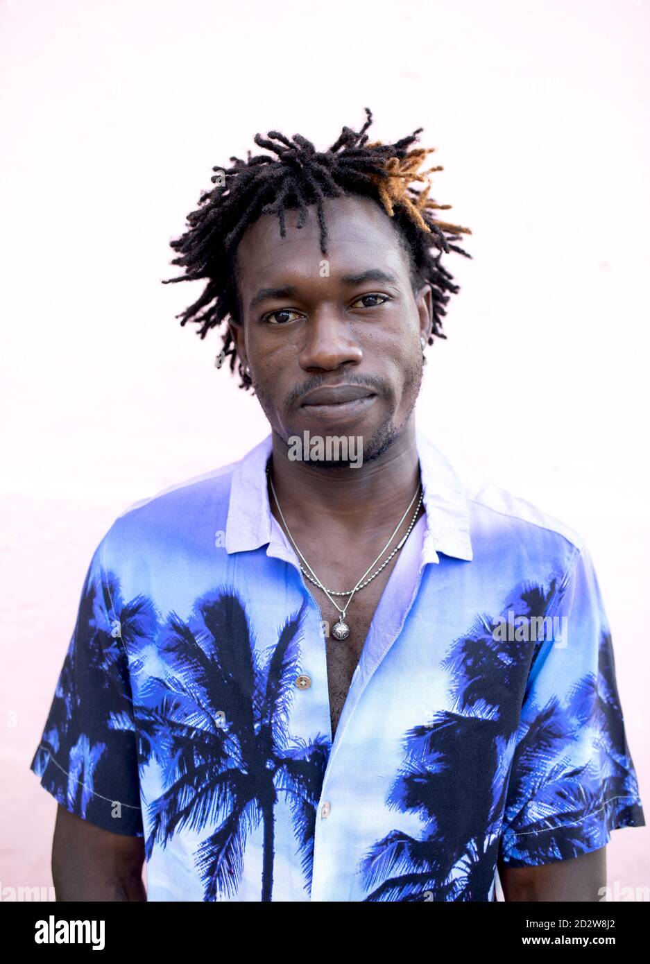 Uomo afro-americano sicuro che indossa una camicia trendy con palme in piedi guardando la macchina fotografica sulla strada vicino all'edificio con rosa parete Foto Stock