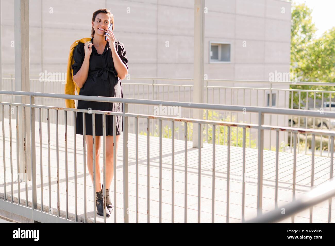 Donna incinta positiva in abito in piedi con giacca sulla spalla e parlare con lo smartphone in una giornata di sole in città Foto Stock
