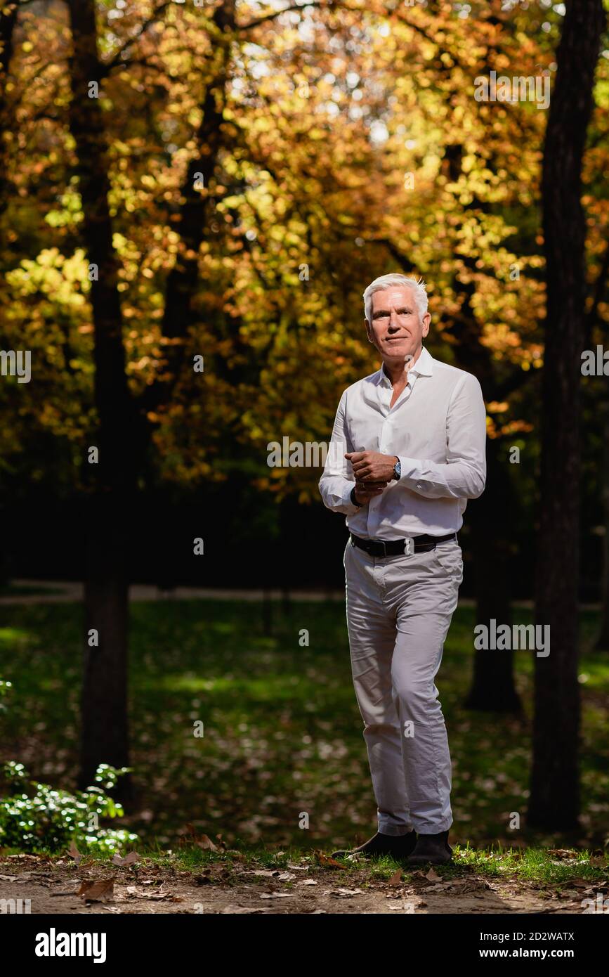 Calmo maschio maturo in elegante vestito in piedi nel giardino della città con alberi gialli e guardando la macchina fotografica Foto Stock