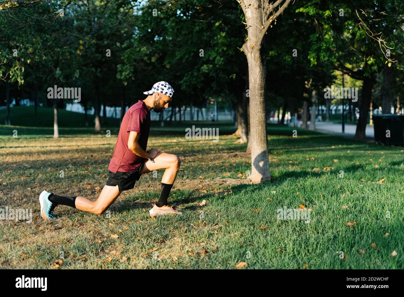 Vista laterale dell'atleta maschile concentrato in abbigliamento sportivo che fa affondo e allungare le gambe mentre si riscaldano durante l'allenamento nel parco Foto Stock