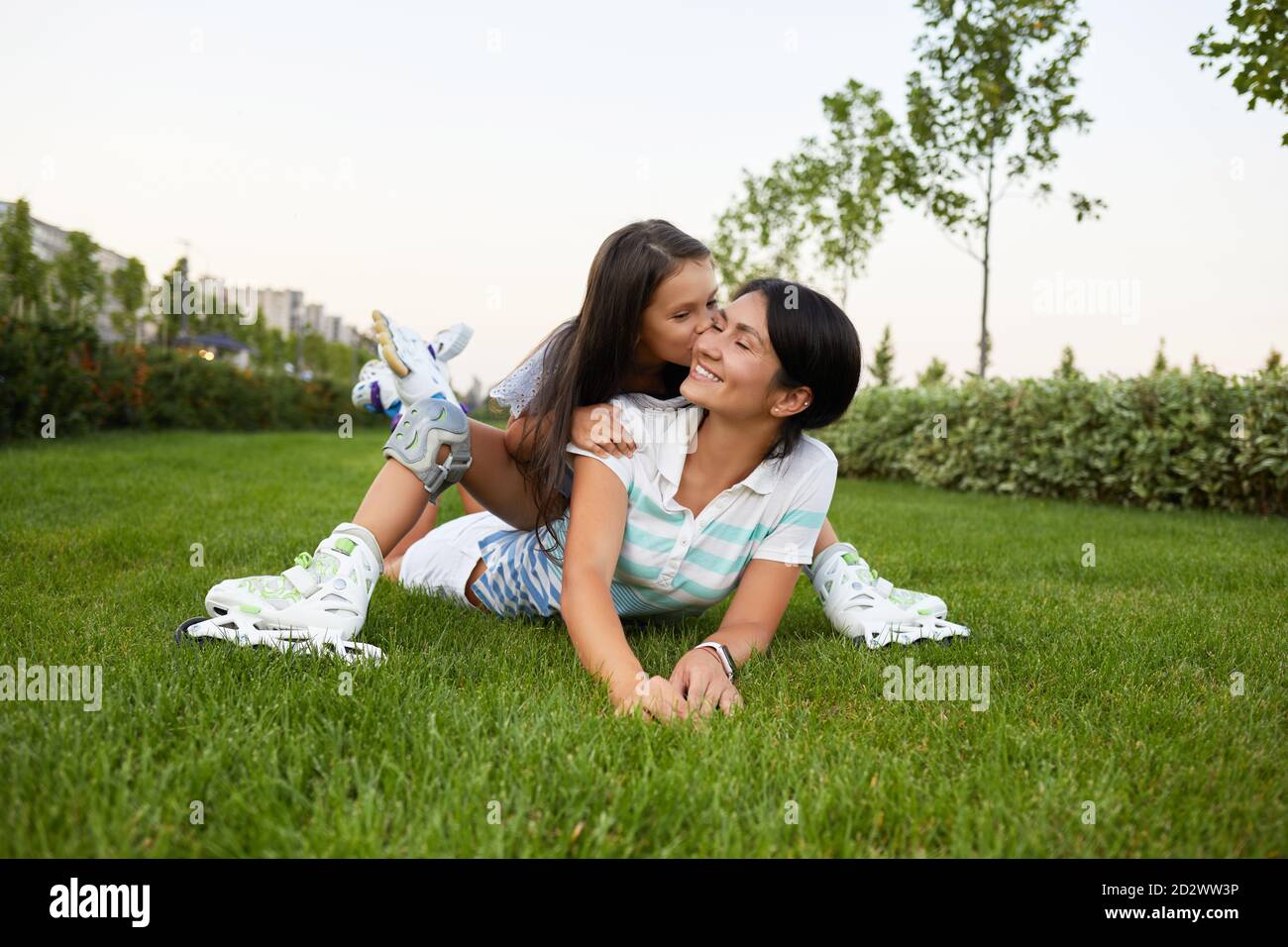 madre felice e figlia in pattini di rullo sono seduti su erba e divertirsi nel parco. bambina sta baciando sua madre Foto Stock