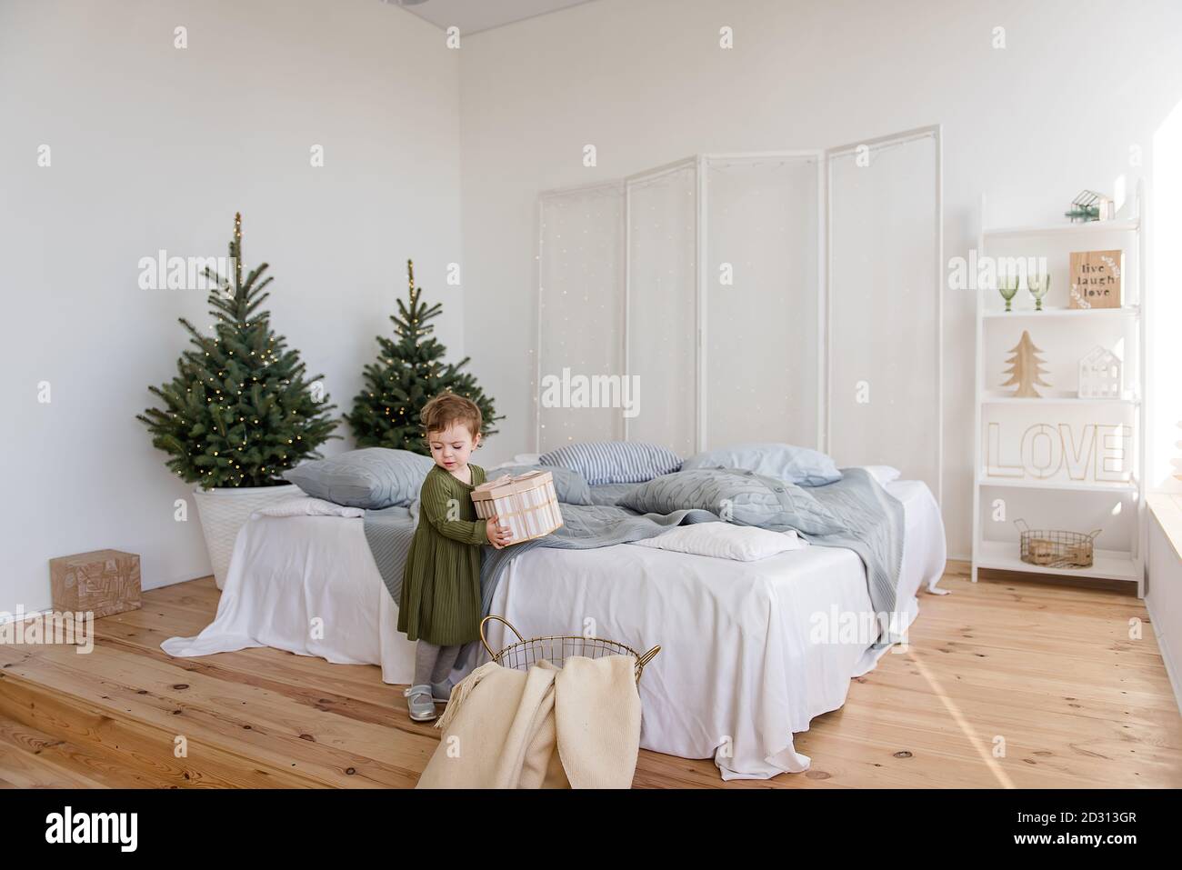 Una bambina in un caldo abito verde ha trovato un regalo da Babbo Natale sul divano. Sfondo in ghirlanda sfocata di luci, albero di Natale. Letto bianco, Foto Stock