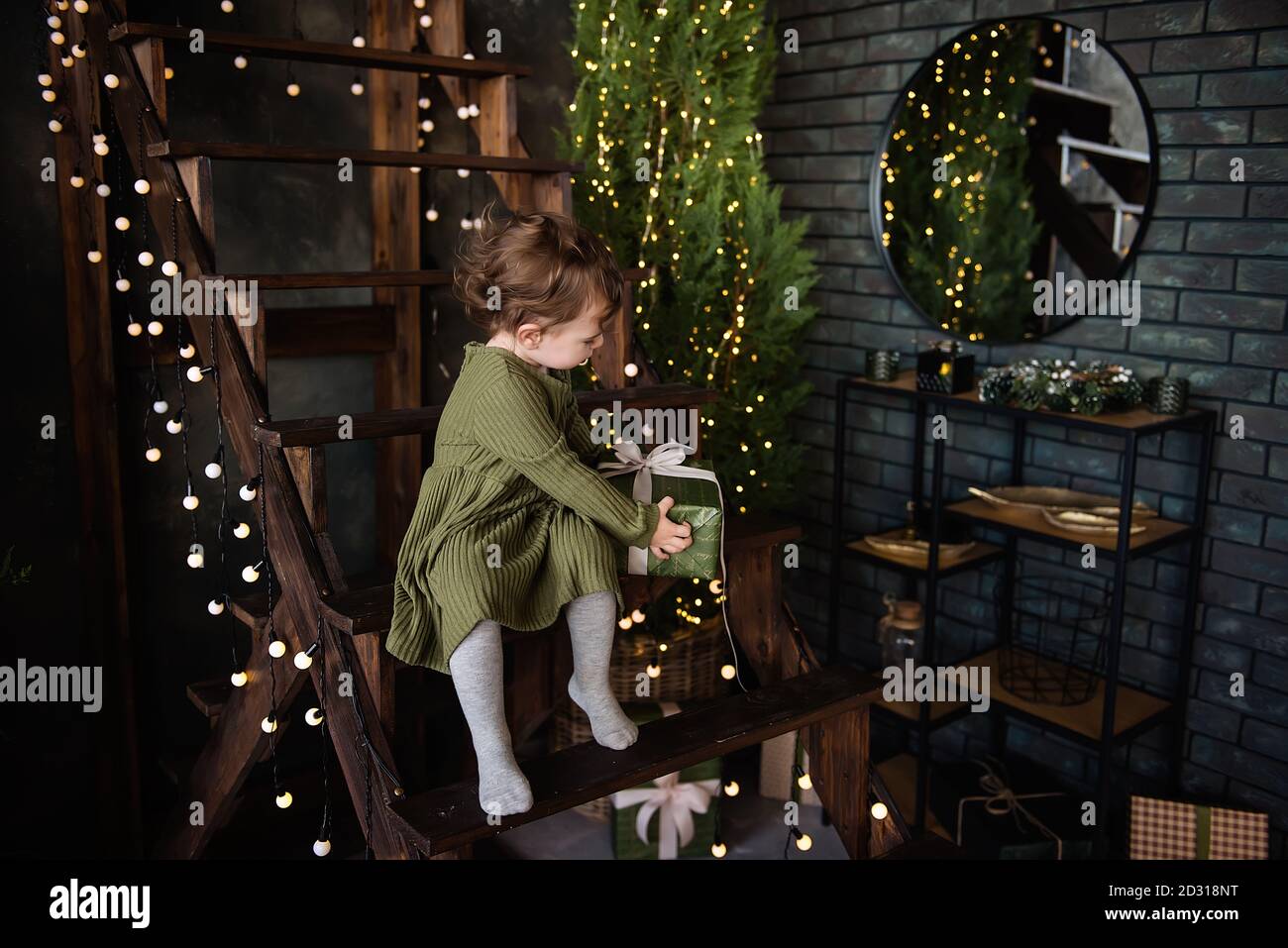 Una bambina in un vestito caldo verde si siede su una scala di legno, tenendo una scatola in un pacchetto con un arco. Regalo da Babbo Natale. Sfondo in luci, Natale Foto Stock
