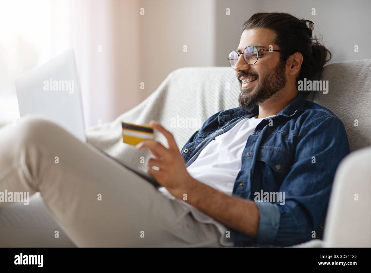 Guy allegro che usa il telefono mobile, la carta di credito ed il laptop che fornisce il pagamento in linea seduto sul lettino a casa. Foto Stock