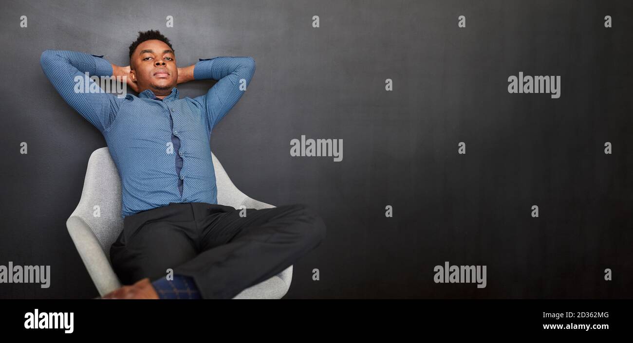 Uomo d'affari africano si appoggia indietro rilassato di fronte ad un grigio muro in ufficio Foto Stock