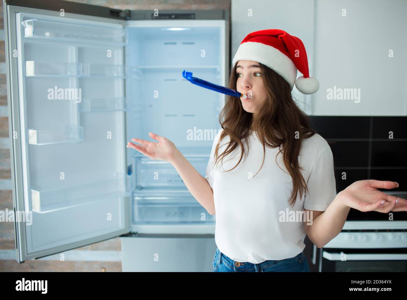 Giovane donna in cucina durante la quarantena. Persona femminile sconvolta in cappello rosso nel periodo di Natale o di nuovo anno. Frigorifero vuoto senza cibo o pasto, fruttato Foto Stock