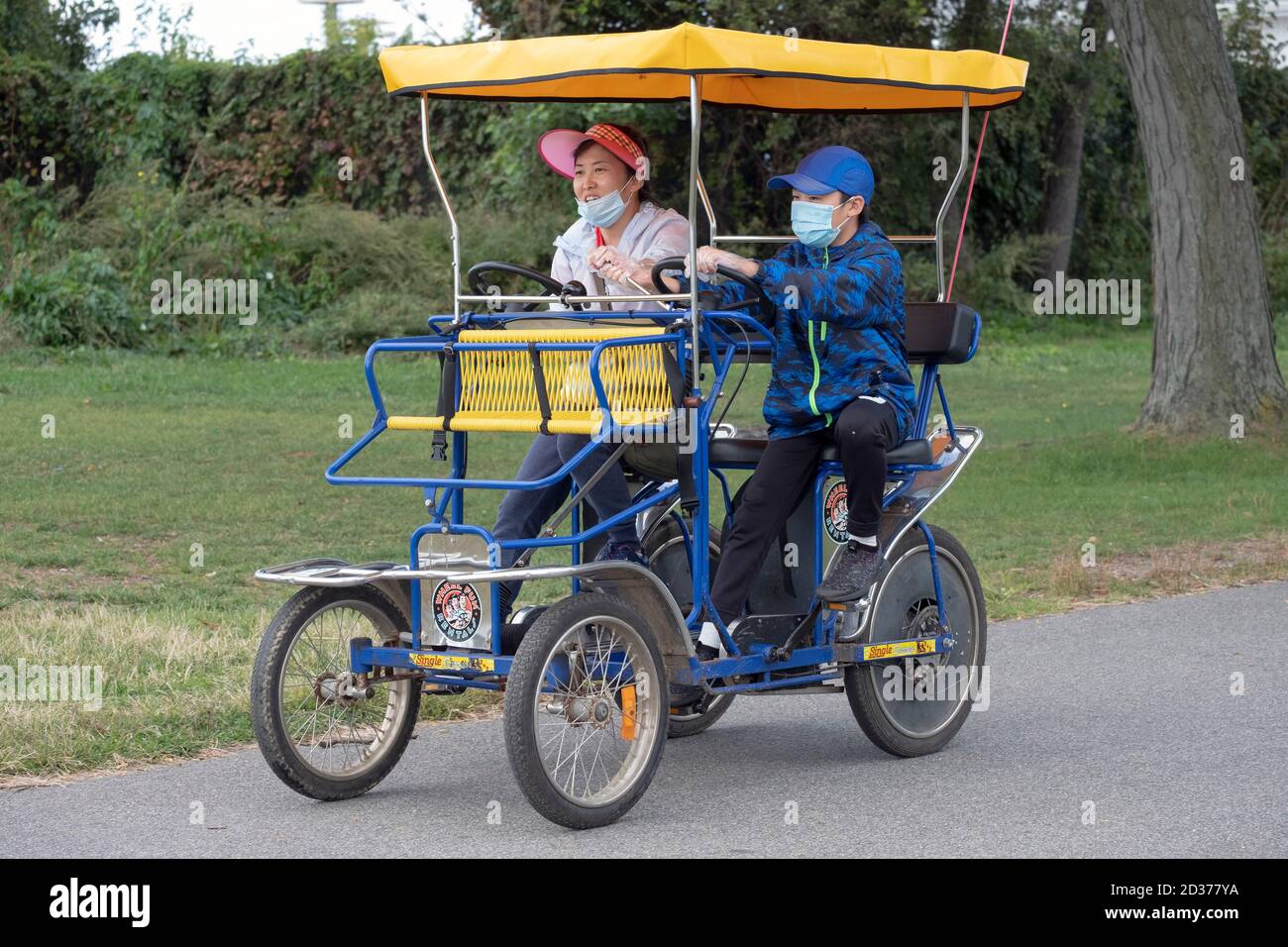 Una madre ed un figlio che indossano le maschere cavalcano un ciclo di surrey a 4 ruote di Divertimento noleggiato. Nel Flushing Meadows Corona Park a Queens, New York City. Foto Stock