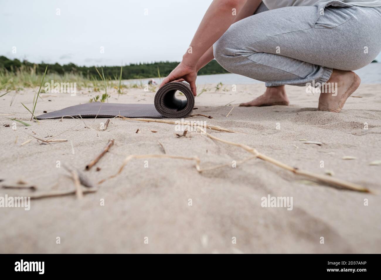 L'uomo dispiega un tappeto da yoga su una spiaggia sabbiosa vicino a un lago, sta per fare esercizi all'aperto in una giornata estiva. Foto Stock