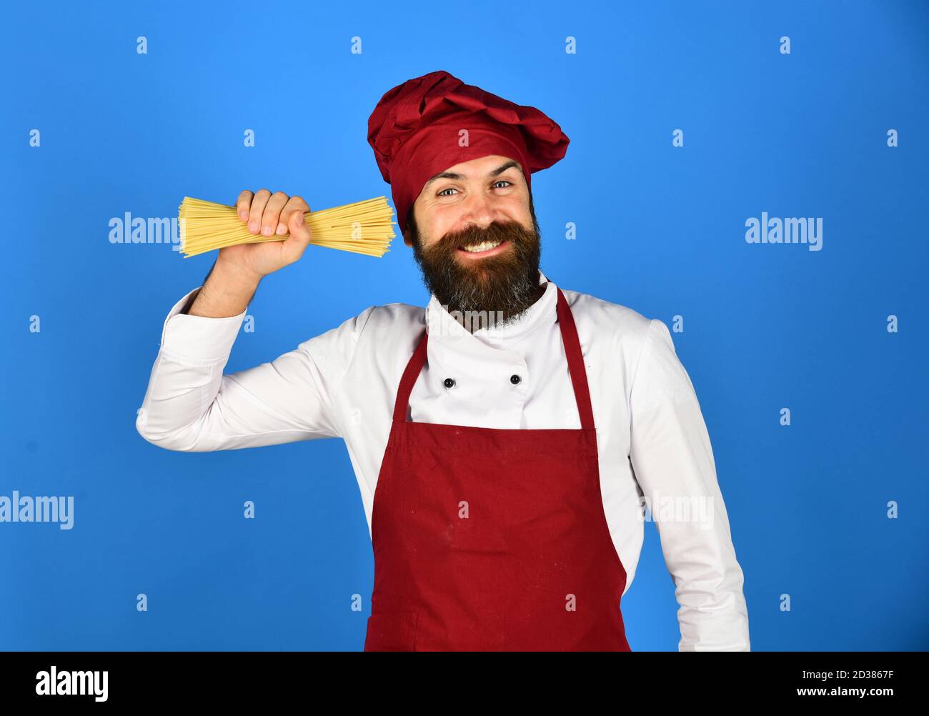 Ristorante italiano. Cuocete con il viso allegro in cappello di borgogna e il grembiule trattiene la pasta secca. L'uomo o l'hipster con la barba tiene i maccheroni vicino all'orecchio su sfondo blu. Chef con mazzo di spaghetti. Foto Stock