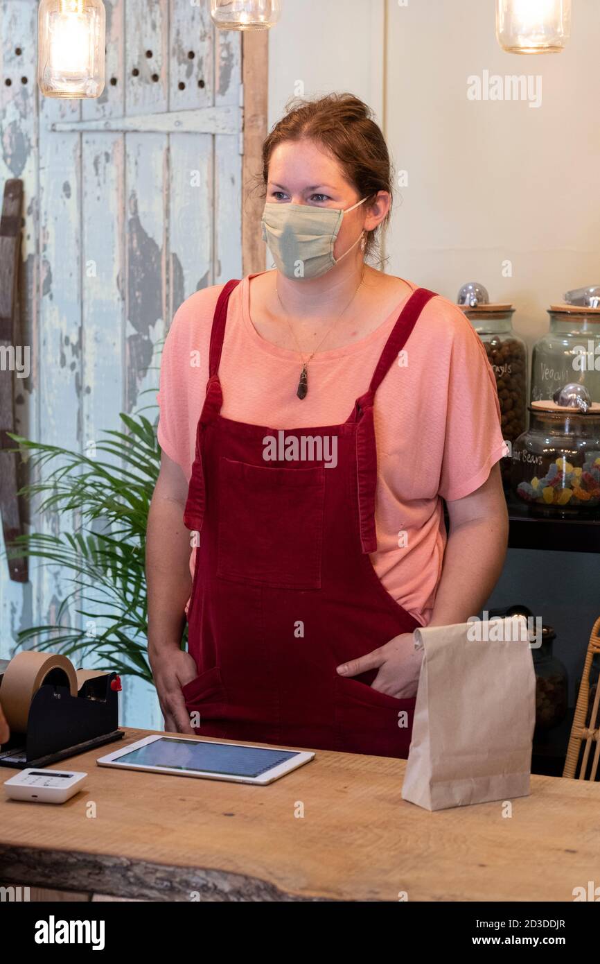 Donna che indossa maschera facciale e grembiule rosso che serve il cliente nel negozio di Wholefood senza sprechi. Foto Stock
