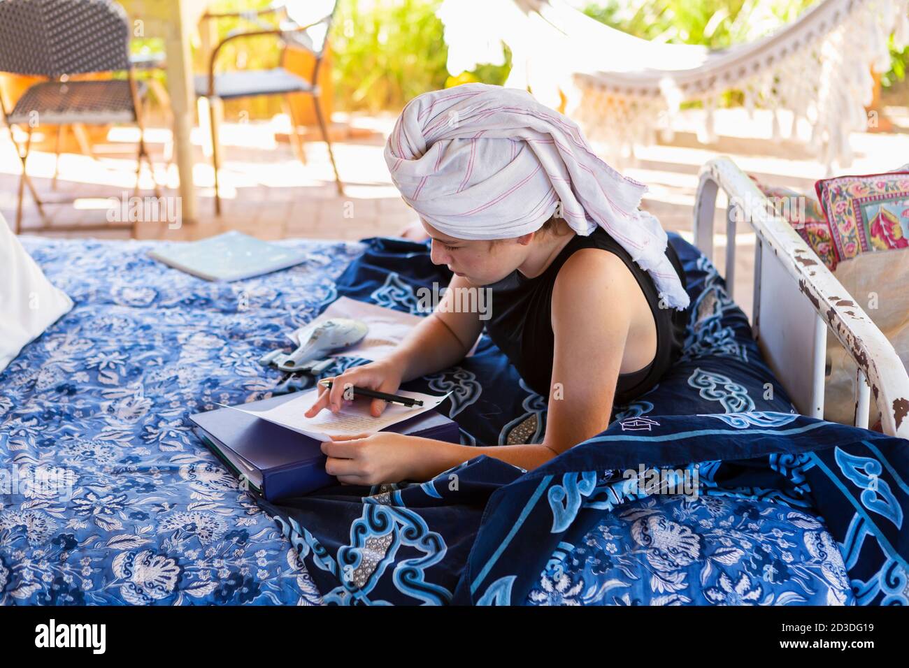 Ragazza adolescente con i capelli avvolti in asciugamano seduto sul letto esterno, facendo i compiti. Foto Stock