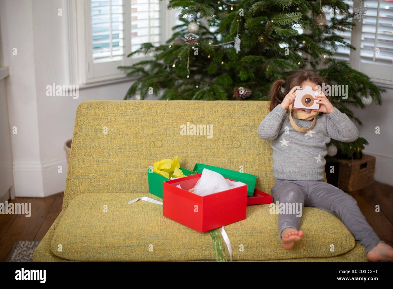 Giovane ragazza seduta sul divano in soggiorno con regalo di Natale in scatola rossa, scattando foto con macchina fotografica giocattolo. Foto Stock