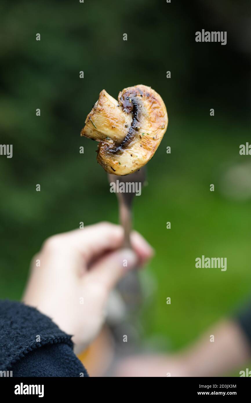 Verdure grigliate all'aperto, una mano femminile che tiene una forchetta con funghi appena grigliati, champignon con erbe. Spuntino vegano. Foto Stock