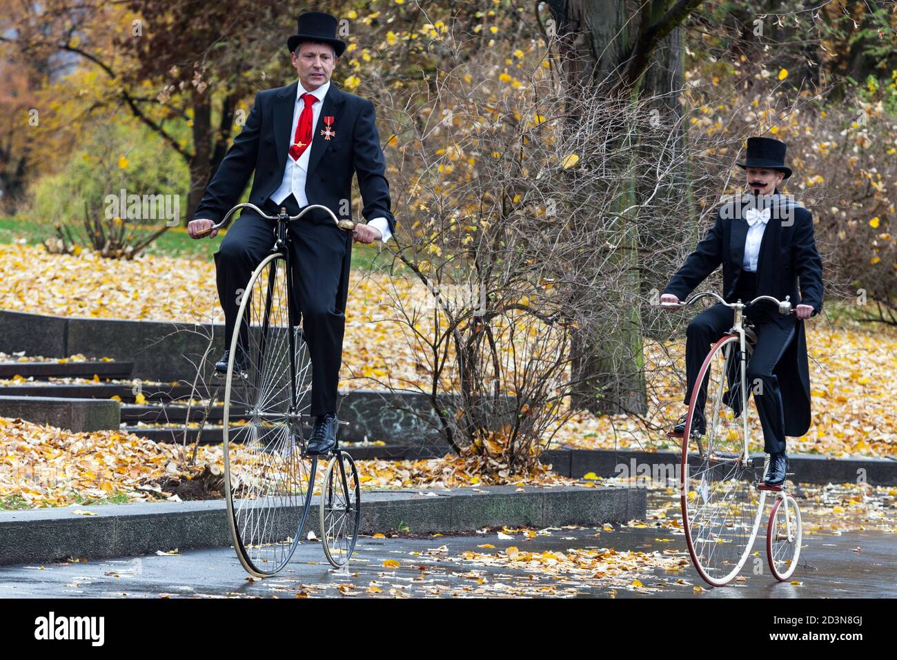 Due ciclisti su biciclette Penny Farthing che indossano un giro sul cappotto di coda attraverso il parco autunnale Foto Stock
