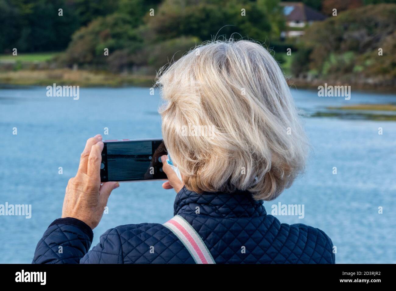 una donna o una donna di mezza età che utilizza un dispositivo cellulare portatile per scattare una foto del mare in vacanza. Foto Stock
