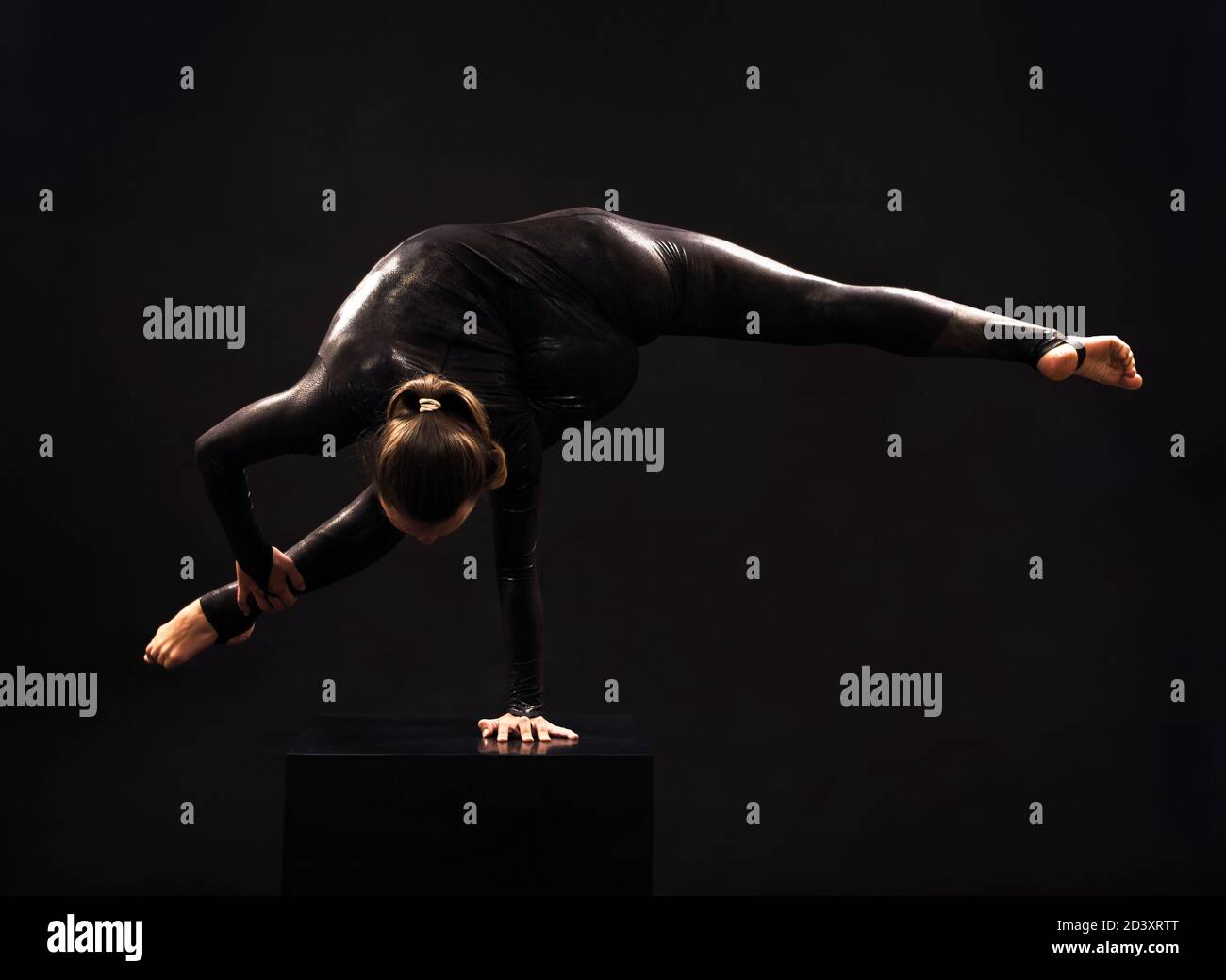 Una donna affascinante ginnastica, in una tuta nera, impegnata in acrobazie alla luce del contra. Immagini isolate su sfondo scuro. Foto Stock