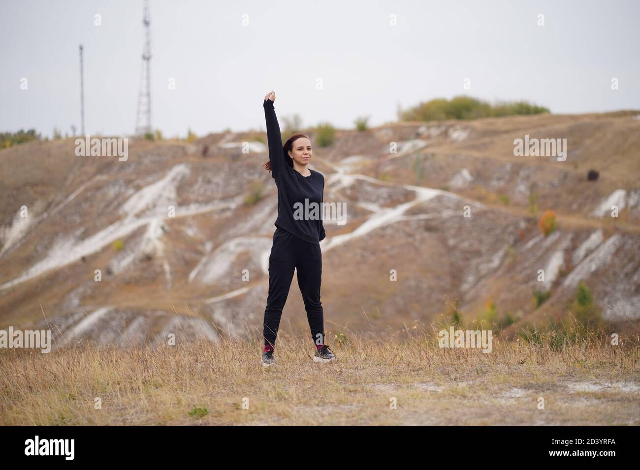 Giovane donna che fa il riscaldamento su terreno collinare. Donna adulta in abiti neri casual facendo sport in aria fresca in campagna. Foto Stock