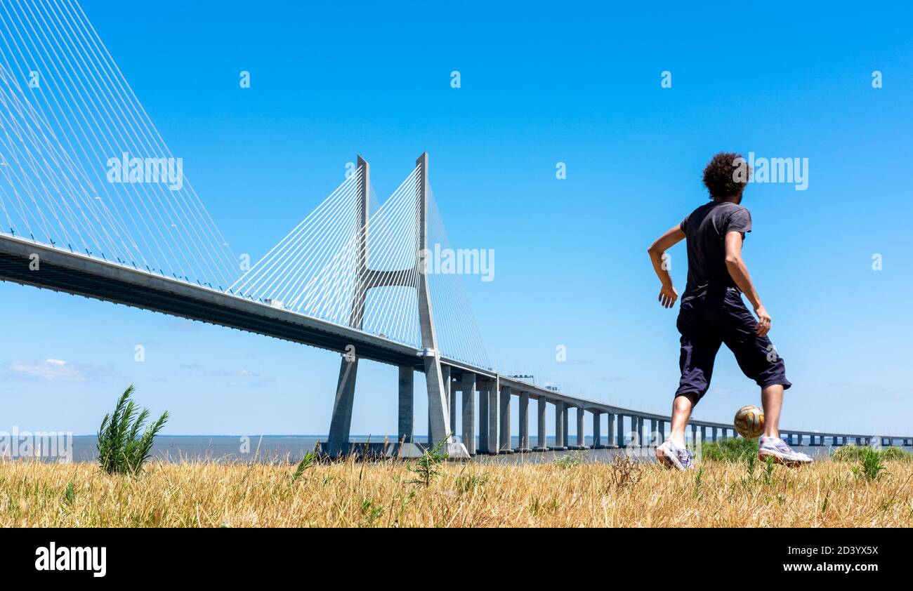 Ragazzo che gioca a calcio in una giornata di sole accanto al ponte Vasco de Gama sul Rio Tejo a Lisbona, Portogallo Foto Stock