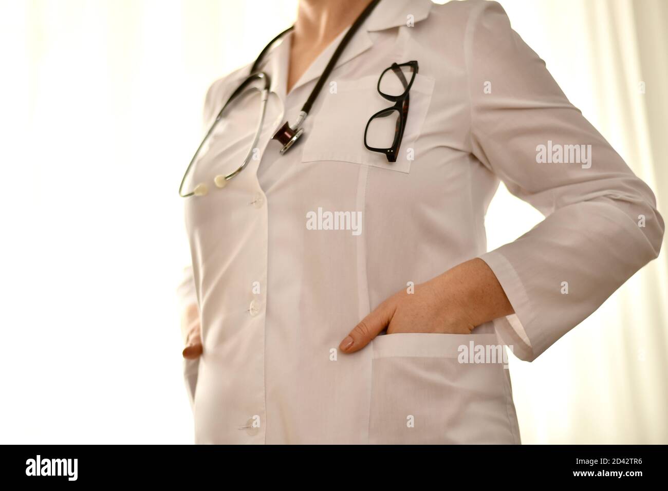 Il medico si alza, entrambe le mani in tasca, uno stetoscopio pende intorno al collo, bicchieri di una cornice scura sul petto. Foto Stock