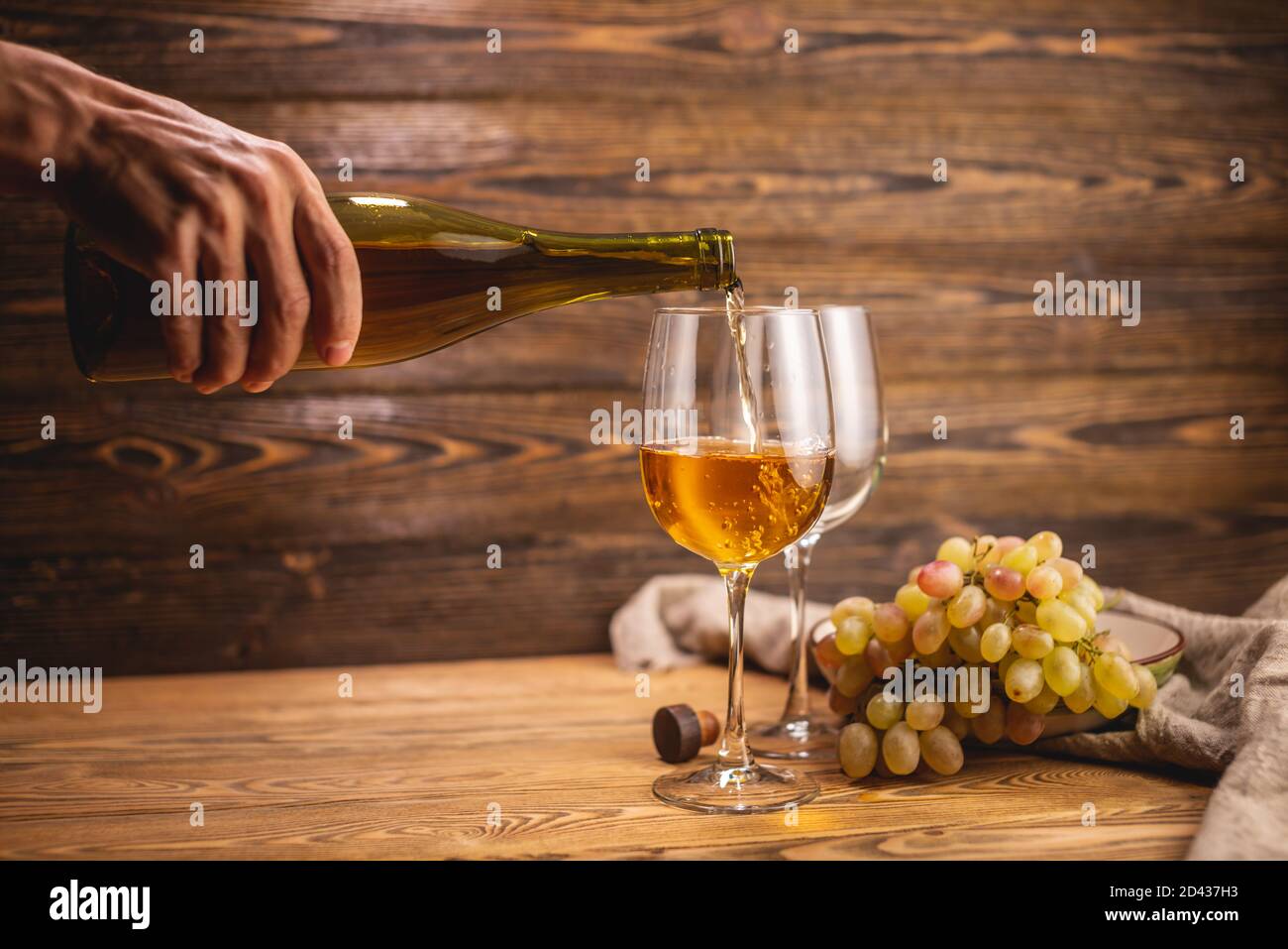 Una mano sta versando vino bianco secco da una bottiglia in un bicchiere sullo sfondo di un mazzo di uva su un tavolo di legno. Concetto di viticoltura e. Foto Stock