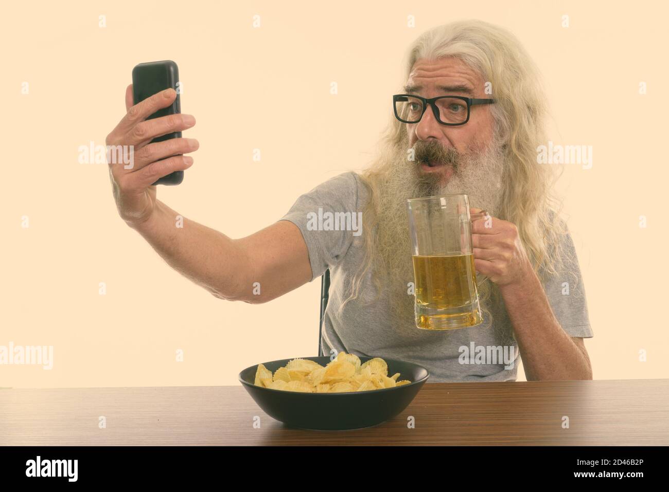 Felice anziano uomo sopportato prendendo selfie mentre tiene il vetro di birra con ciotola di patatine su tavolo di legno Foto Stock