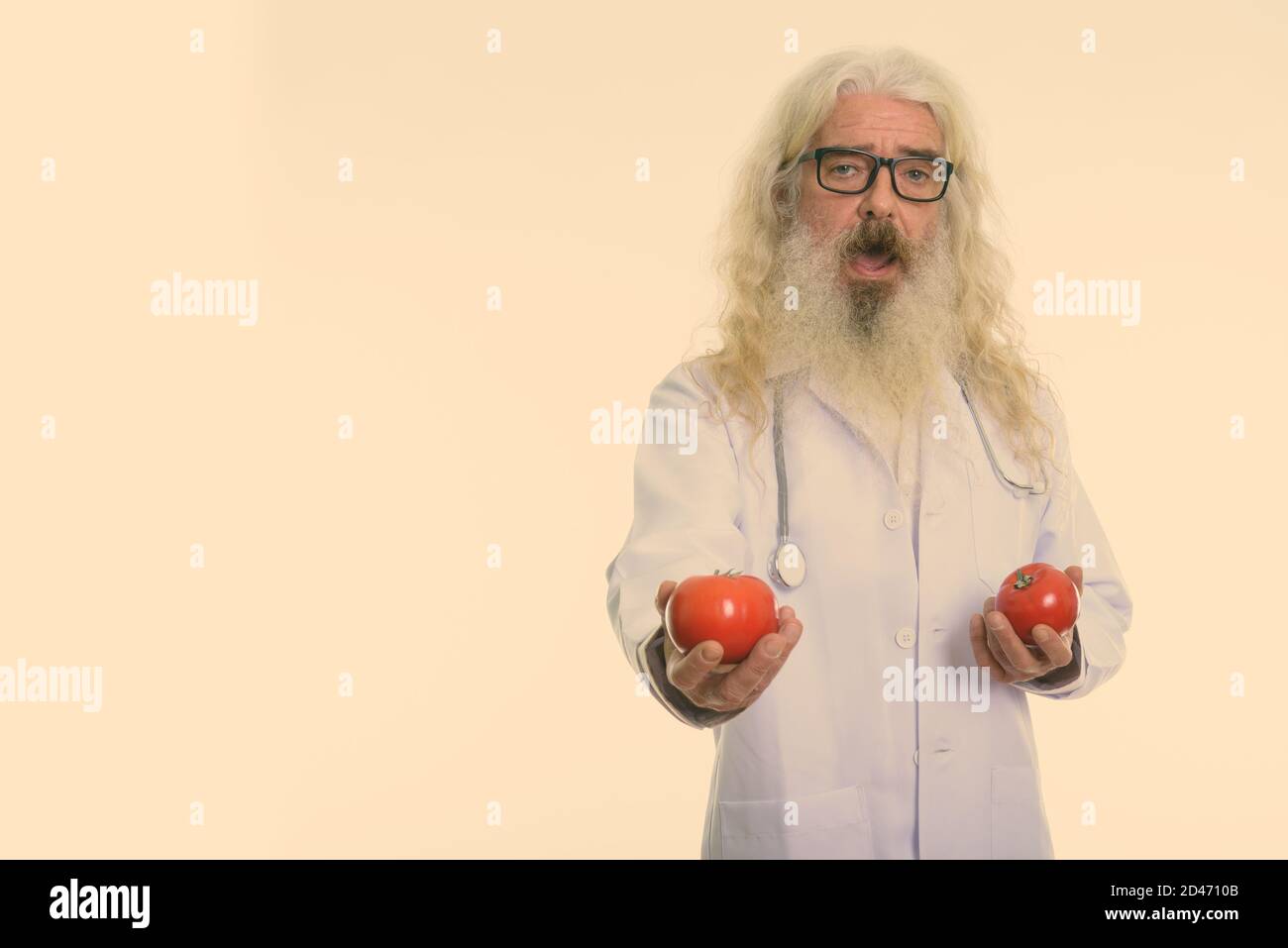 Studio shot di felice senior uomo barbuto medico sorridente tenendo premuto e dando pomodori rossi Foto Stock