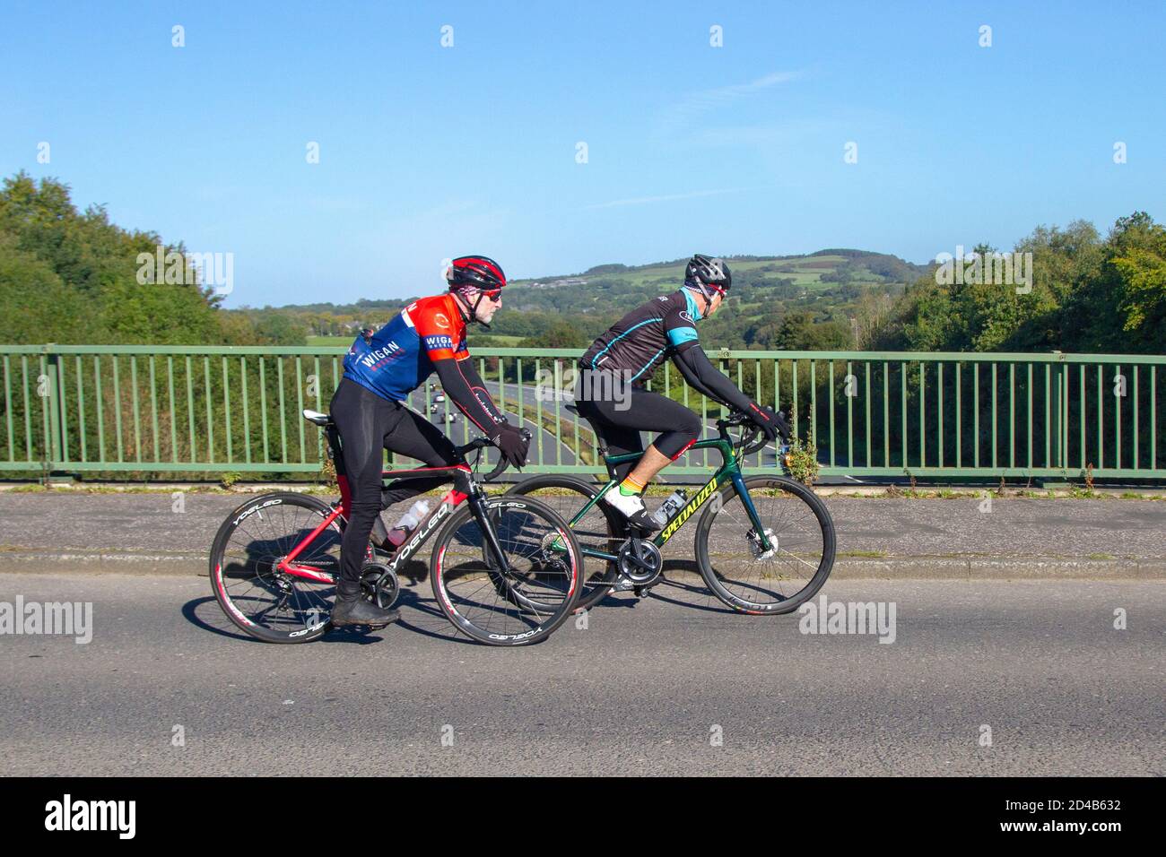 Due ciclisti maschi Wigan che guidano specializzati & Yoeleo sport bici da strada su strada di campagna che attraversa il ponte autostradale nella rurale Lancashire, Regno Unito Foto Stock